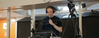 A man stands behind a soun desk wearing headphones, talking into a microphone. Beside him is a camera pointing to the front of the venue.