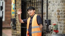 A man in an orange jacket waves while holding a gate open