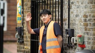 A man in an orange jacket waves while holding a gate open