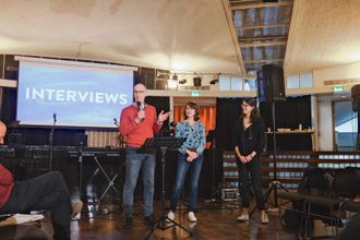 Three people stand at the front of church; two of them are being interviewed by the third.