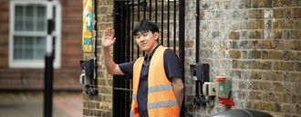 A man in an orange jacket waves while holding a gate open