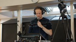 A man stands behind a soun desk wearing headphones, talking into a microphone. Beside him is a camera pointing to the front of the venue.