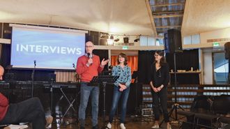 Three people stand at the front of church; two of them are being interviewed by the third.