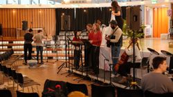 A group of musicians practicing at the front of a school hall. There is a keyboard, guitar, cello, and drumkit.