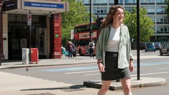 A London scene, outside Elephant and Castle tube station. There is a red bus in the background. A person is crossing the street from the tube station walking towards the city.