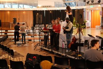 A group of musicians practicing at the front of a school hall. There is a keyboard, guitar, cello, and drumkit.